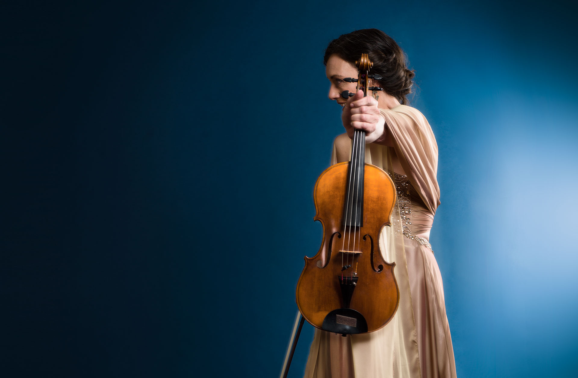 Violinist i guldig klänning håller fram fiolen mot kameran, bakgrunden är blå.