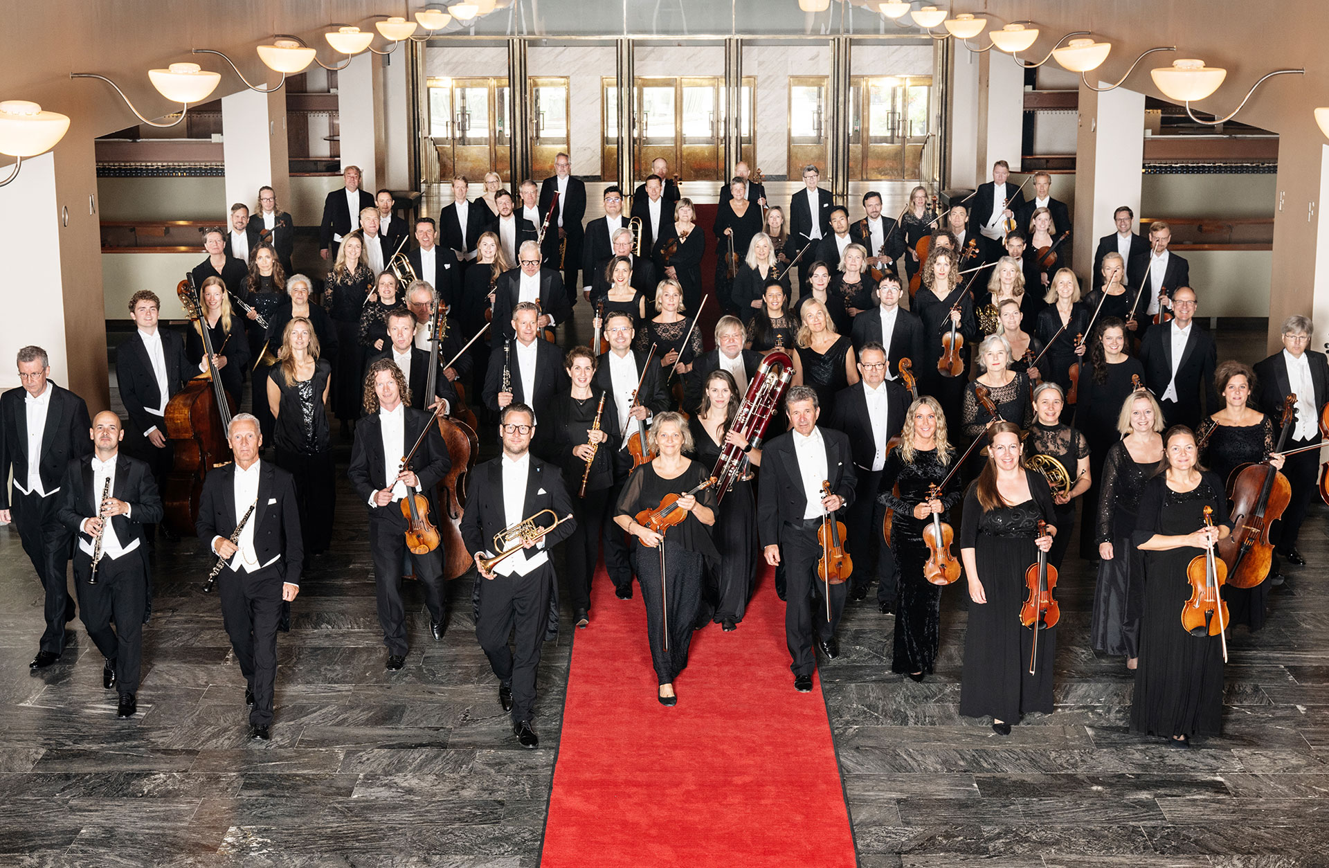 Göteborgs Symfoniker går mot kameran med instrumenten i handen, på en röd matta.