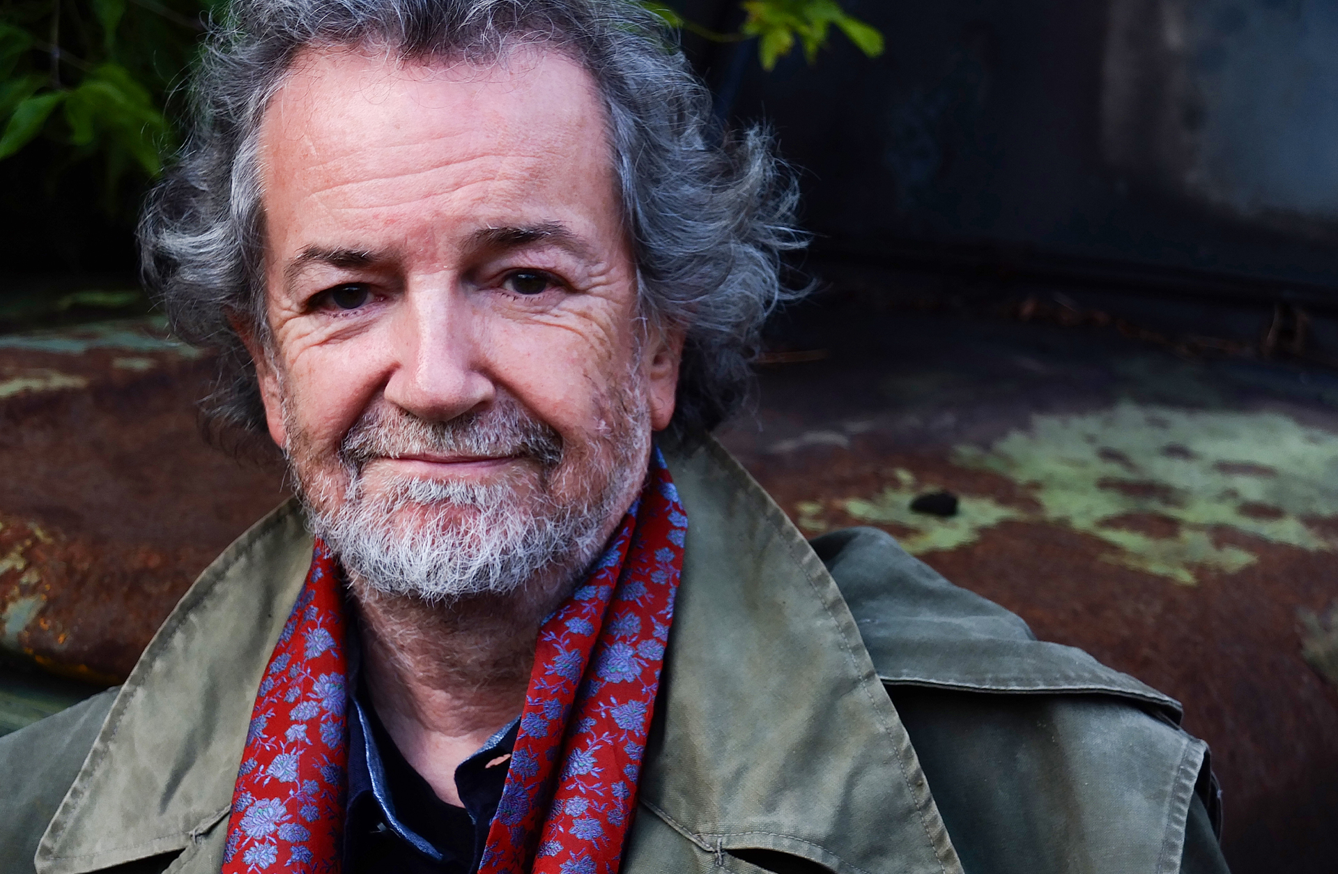 Porträtt av Andy Irvine, en äldre man med vänligt leende. Han har grått hår och skägg och ler mot kameran.