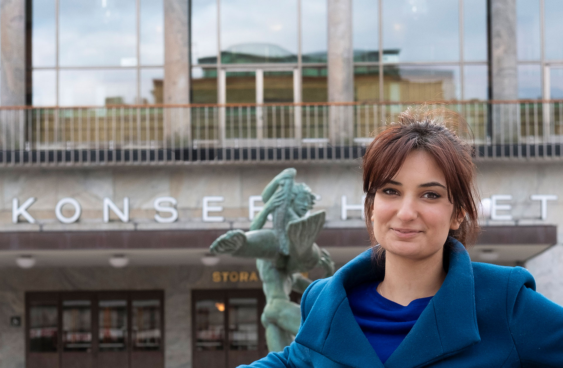 Ava Bahari står utanför Göteborgs Konserthus, vid fontänen Poseidon. Hon har en blå ullkappa och hon ler mot kameran.