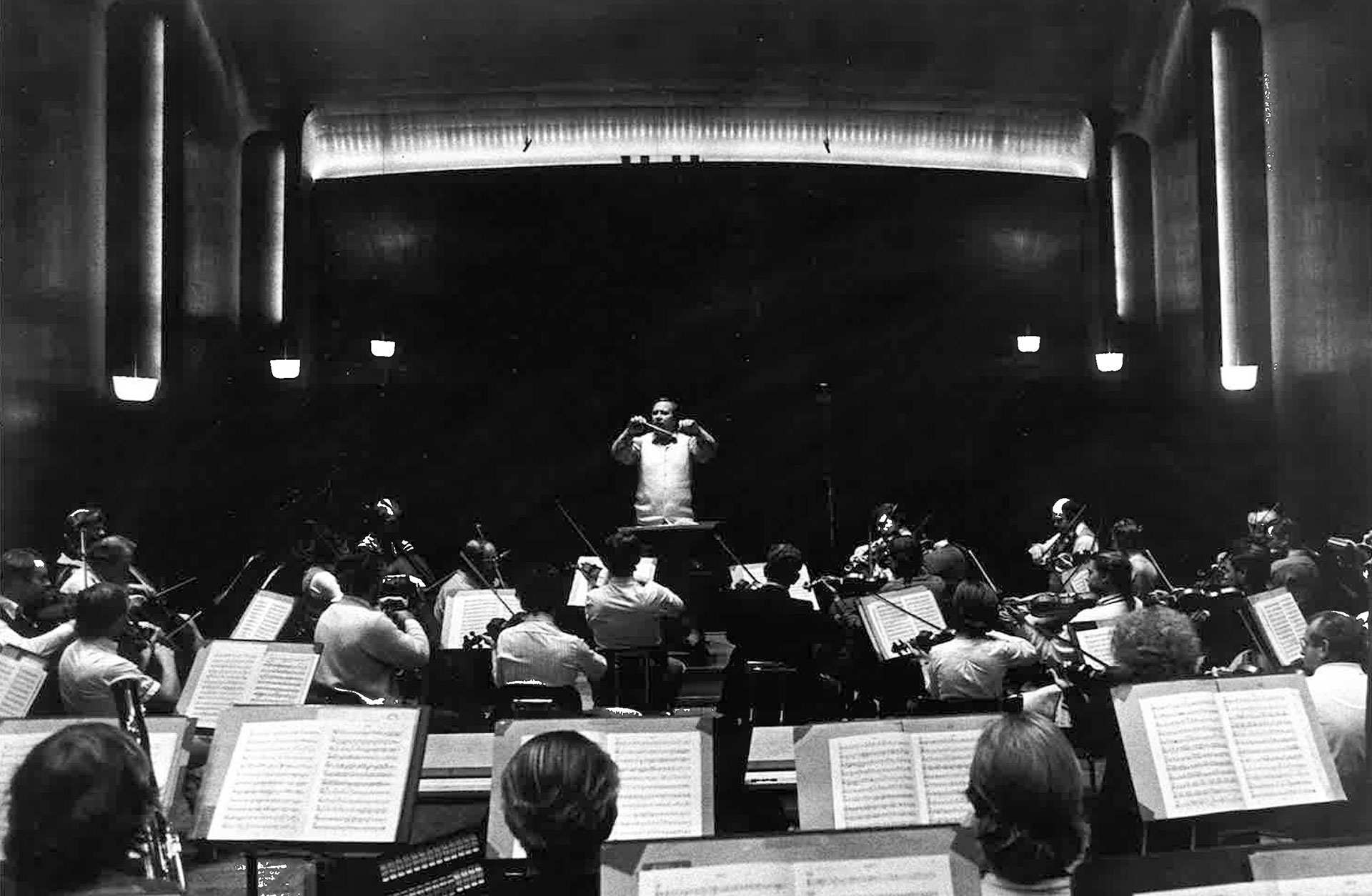 Svartvit bild från 1980-talet där Neeme Järvi står på pulten framför Symfonikerna i Göteborgs Konserthus.