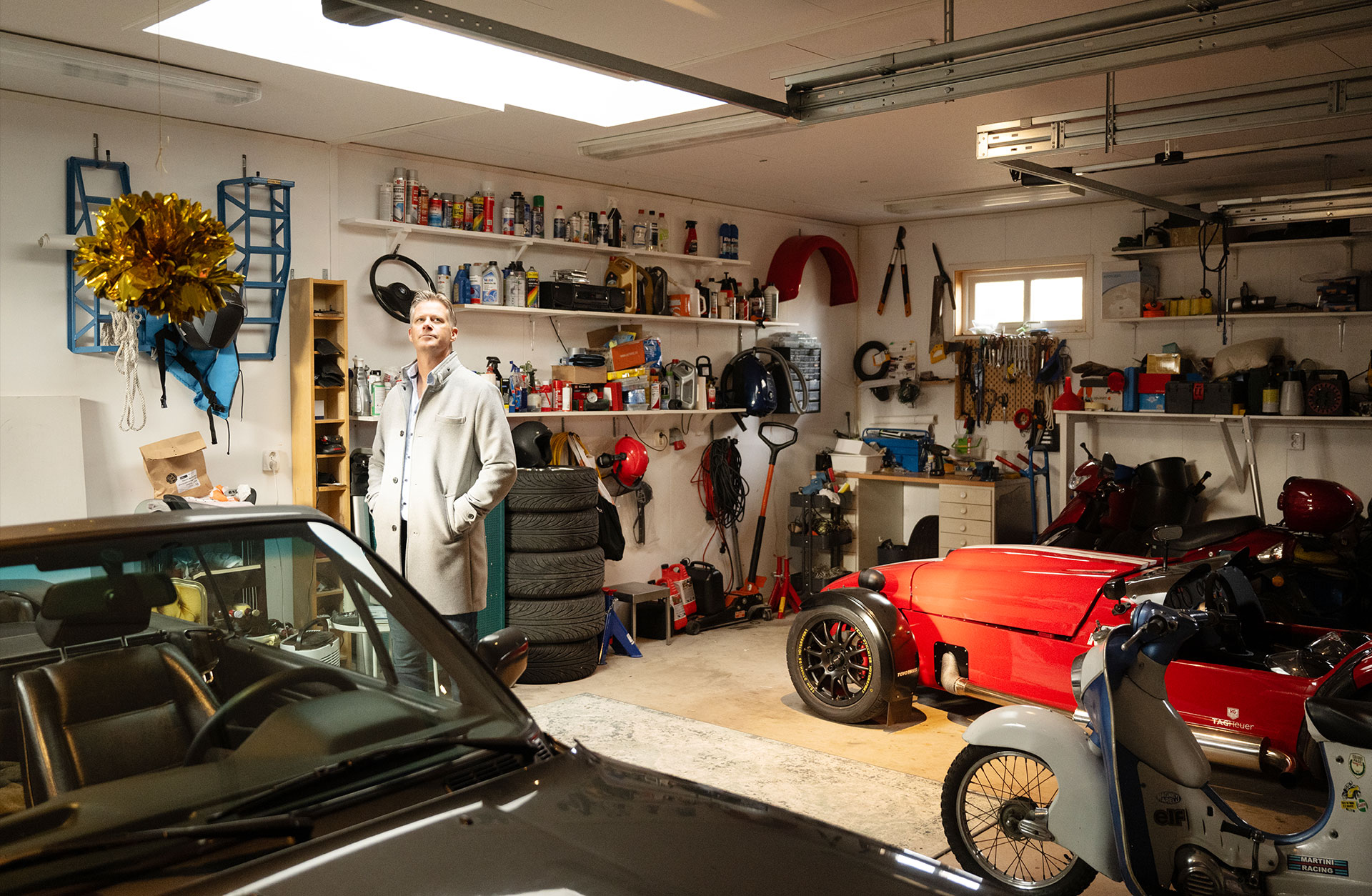 Henrik Nordkvist står i sitt garage, vid en bil och en vespa.