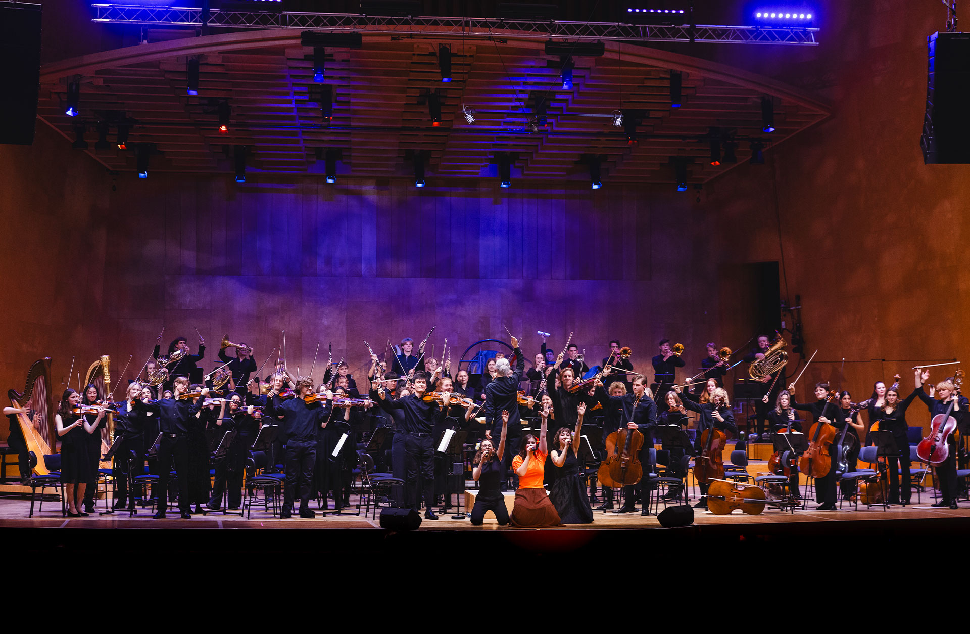 Vägus-orkestern befinner sig på Konserthusets scen. Musikerna står upp med armar och instrument i luften. De är engagerade och spelar.